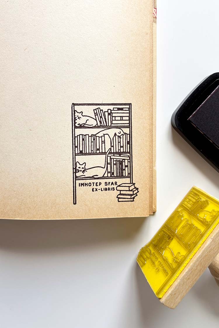 Portadilla de libro estampada con una estantaría llena de libros y tres gatos y sello