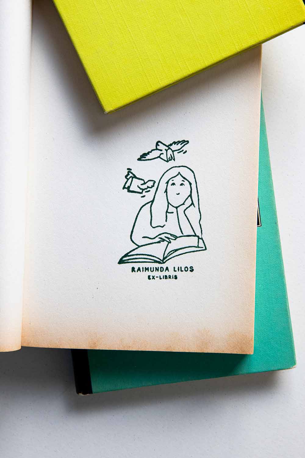 Portadilla de libro estampada con sello exlibris de una mujer leyendo un libro, con dos libros volando sobre ella