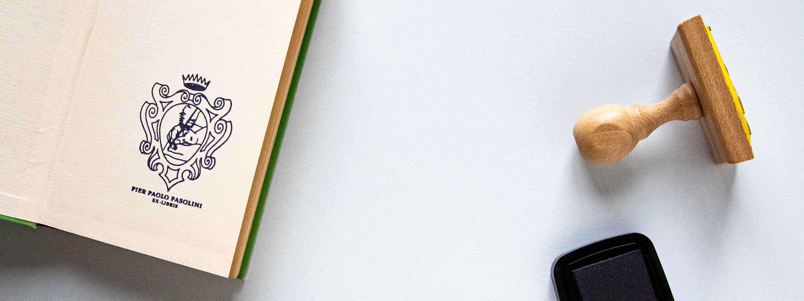 Exlibris personalizzato timbrato sulla pagina di copertina di un libro aperto con uno scudo araldico con una corona, all’interno del quale si vede una mano che scrive con una penna d’oca